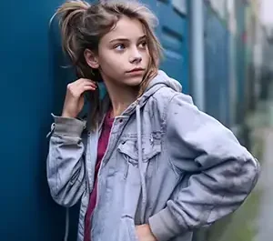 Jeune adolescente de profil en veste à capuche grise avec une main dans les cheveux, posant devant un mur bleu dans un style streetwear urbain