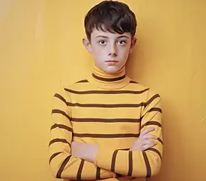 Portrait candide d'un jeune garçon roux au regard étonné, portant un haut rayé jaune et gris devant un mur jaune vif, avec des ombres graphiques.