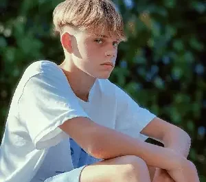 Grand ado assis sur un banc avec une attitude boudeuse et un regard pensif, portant un t-shirt blanc sous une lumière vive en extérieur.