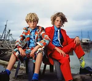 Deux adolescents, costumes colorés (rouge et motifs bleus) assis dans une déchetterie navale, créant un fort contraste visuel avec les débris de bois.