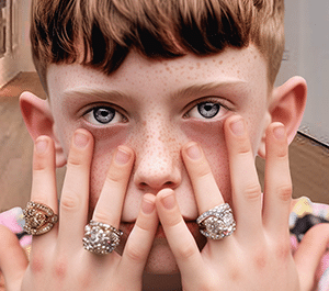 Jeune garçon en galerie d’art, mains devant le visage avec bagues, pose artistique et style affirmé.