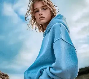 Jeune éphèbe aux cheveux blonds légèrement mouillés, vêtu d’un hoodie bleu et d’un pantalon beige, posant sur des rochers en bord de mer sous un ciel nuageux.