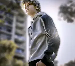 Jeune garçon aux cheveux blonds en silhouette, portant un sweat à capuche gris et noir, posant avec les mains dans les poches sous un éclairage en contre-jour.